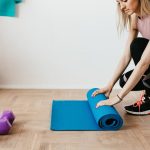 Side view of young female athlete in sportswear unfolding blue sport mat and looking down while sitting on floor near dumbbells and water bottle preparing for training indoors